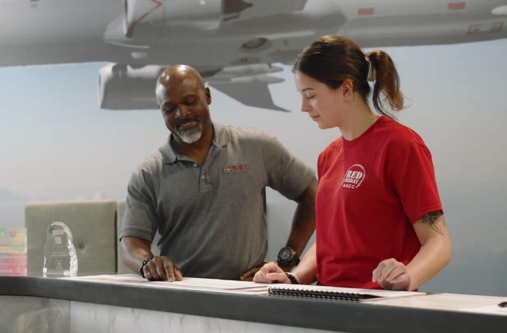 Two people sign documents at a counter: a man in a gray polo and a woman in a red shirt, with an airplane model hanging above them.