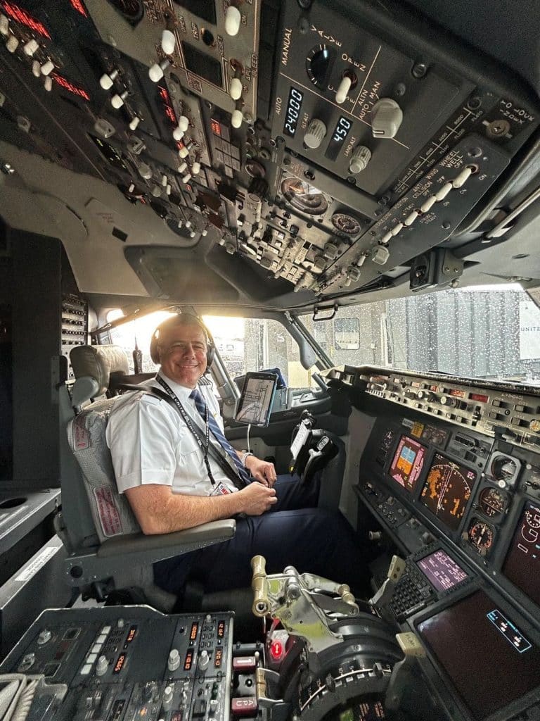 Smiling pilot in uniform sits in the cockpit surrounded by flight panels, gauges, and switches.