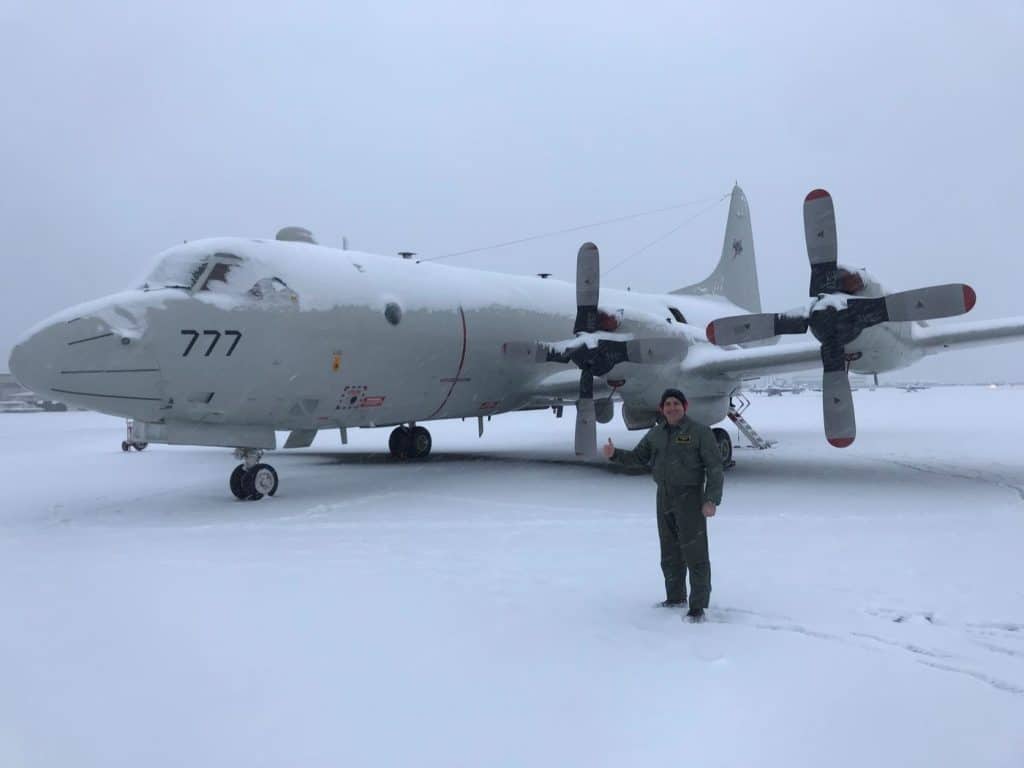 Military transport aircraft with four propellers parked on a snow-covered tarmac; crew member in green flight suit stands in front giving a thumbs-up beside nose number 777.