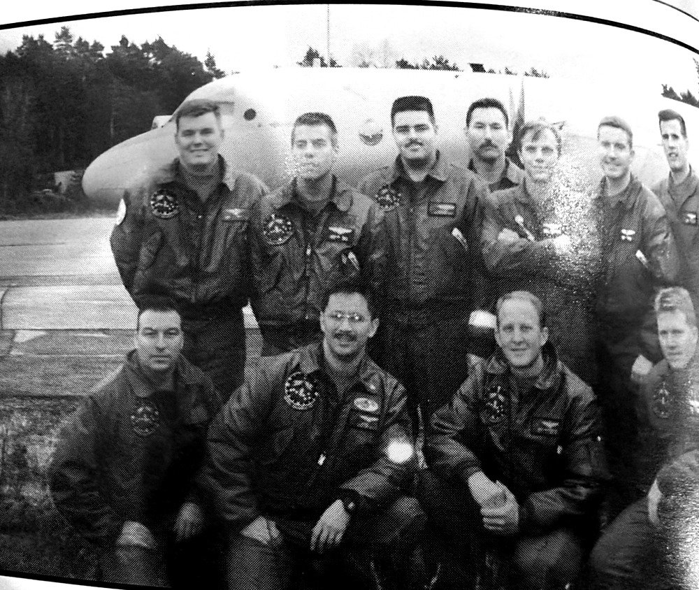 Group of astronauts in matching flight suits posing in front of a large airplane on an airfield/ runway. They wear patches on their jackets, suggesting a spaceflight team photo.