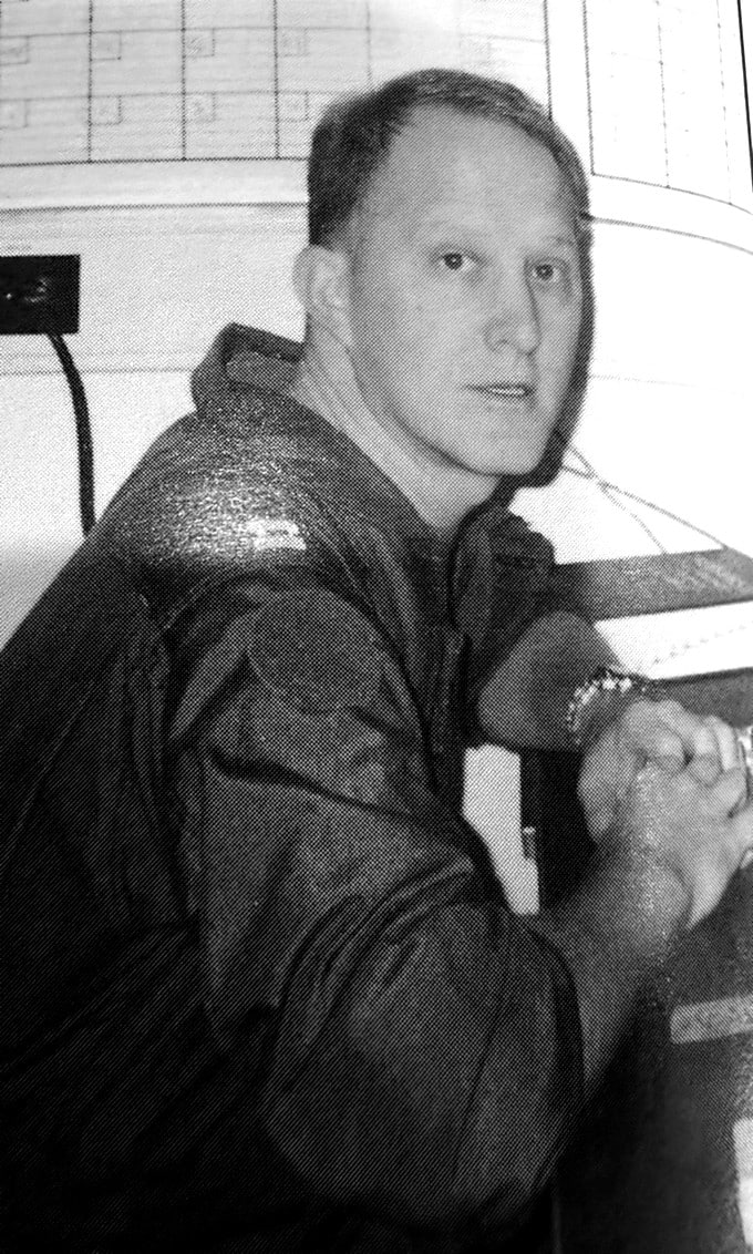 Man wearing a headset sits at a desk in a control room, looking toward the camera.