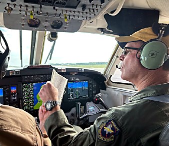 Pilot wearing a green uniform and headset in the cockpit, reviewing a paper checklist at the controls with instrument panels and windows in view
