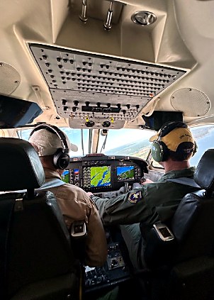Pilot and co-pilot wearing headsets sit in a small airplane cockpit, with navigation displays and overhead controls visible.